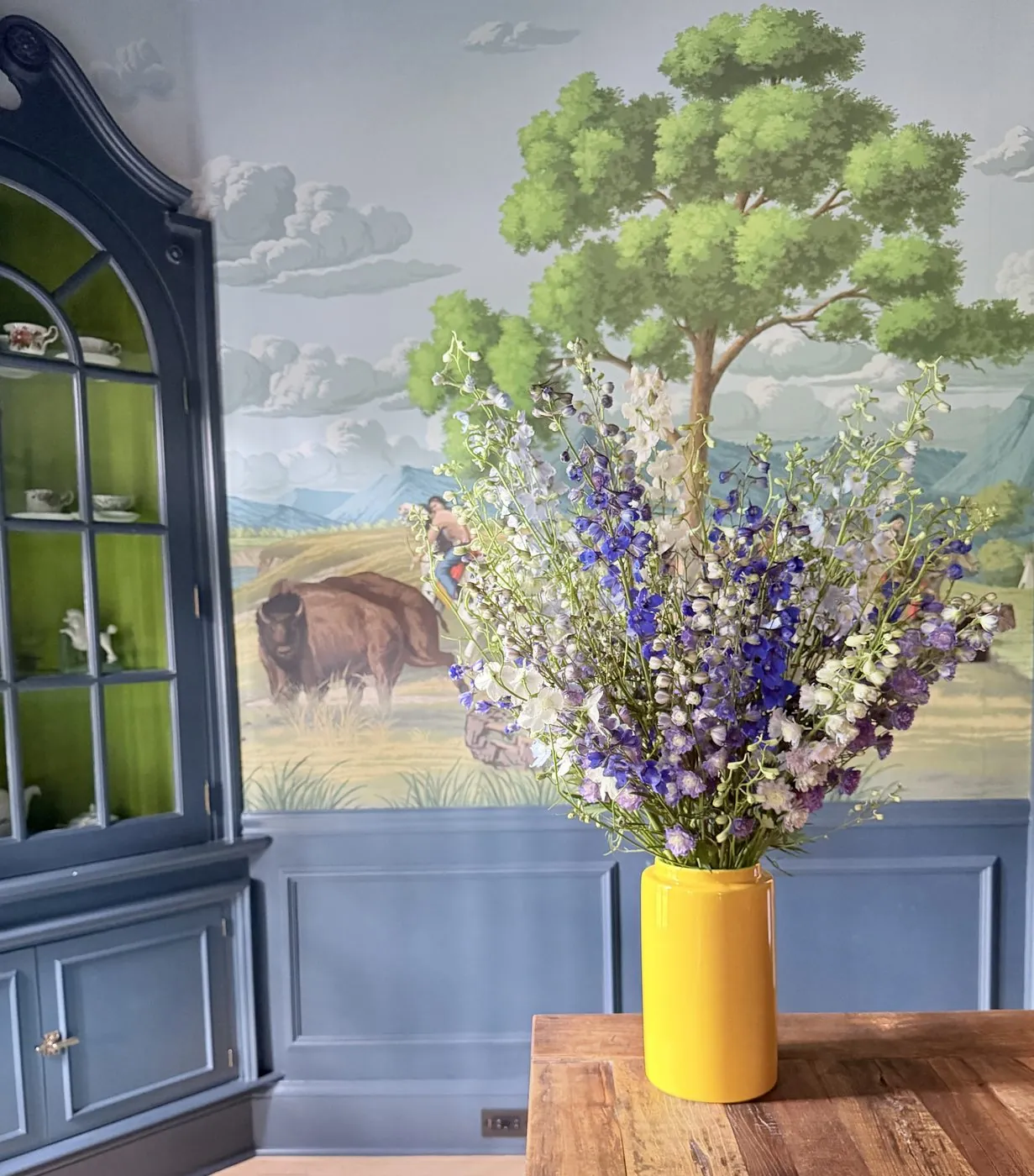 Delphinium and larkspur in a yellow vase before a prairie mural